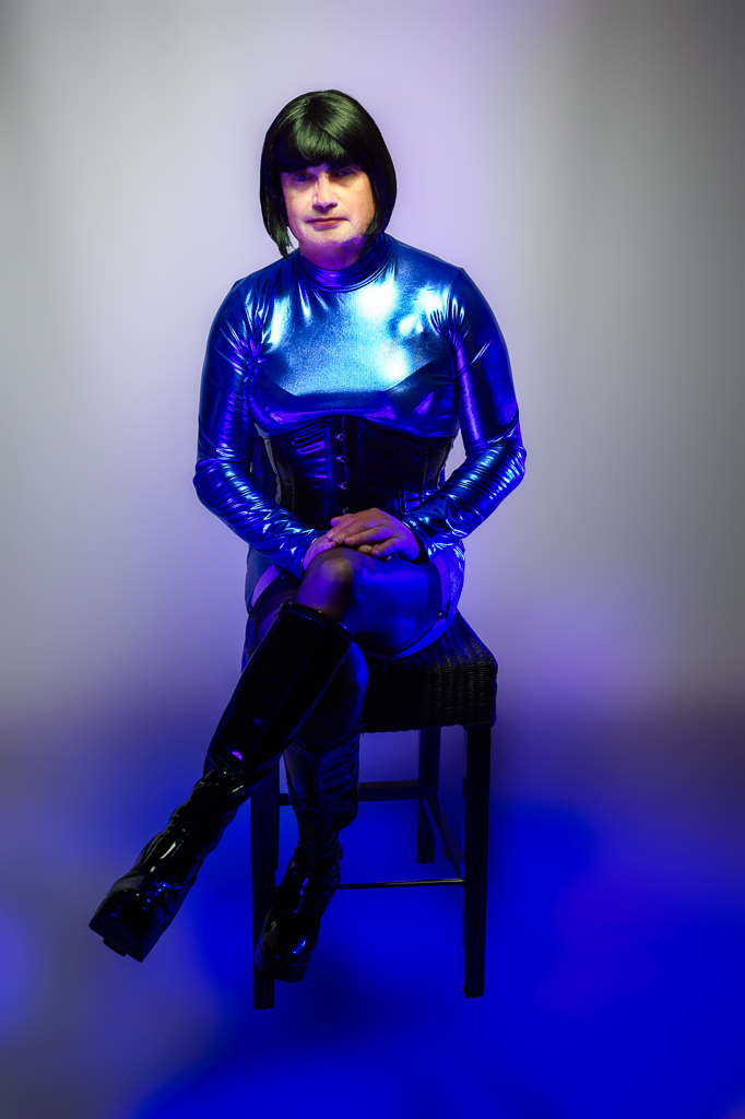 Maria seated on a high stool in a shiny blue dress, studio, blue/purple lighting, full body