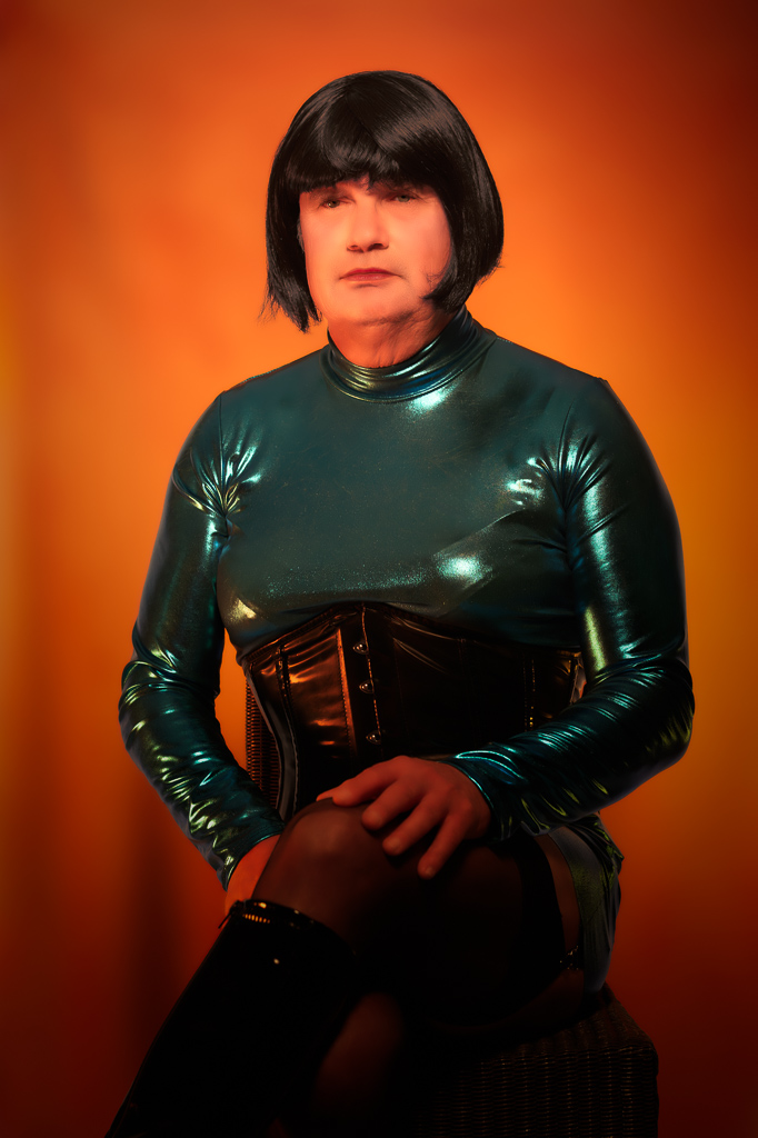 Maria seated on a high stool in a shiny blue dress, studio, waist-up