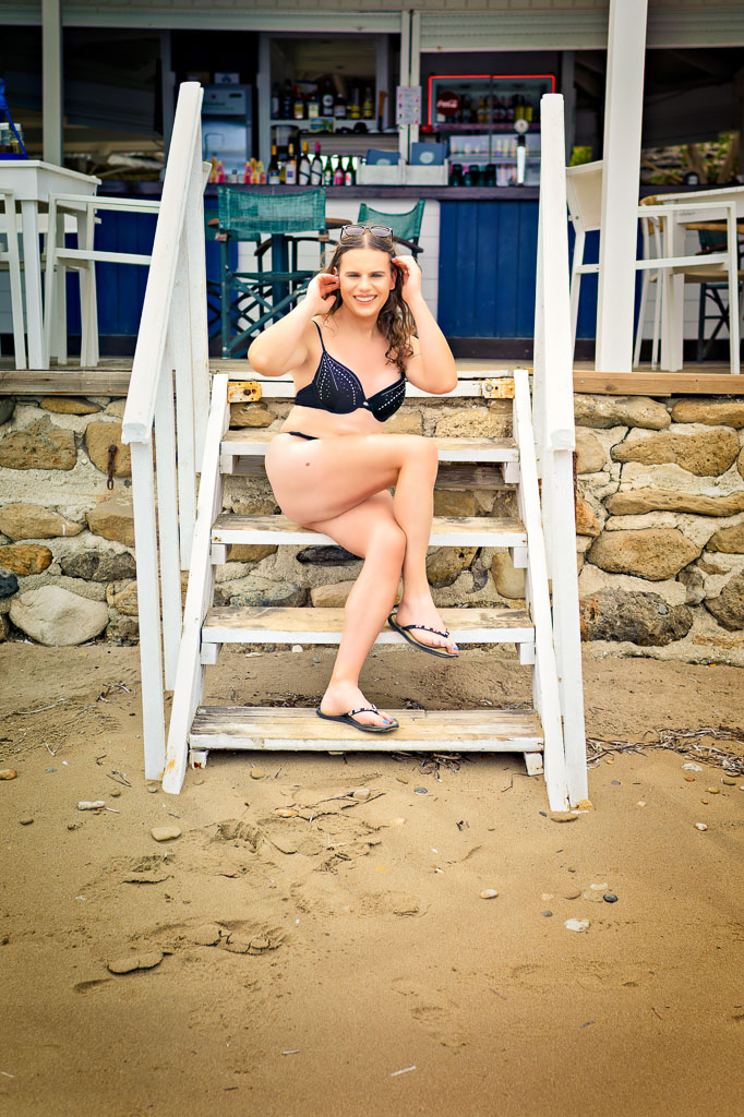 Jessica in a black swimsuit, sitting on the white steps up to the beach bar, Kaminia beach, Zakynthos