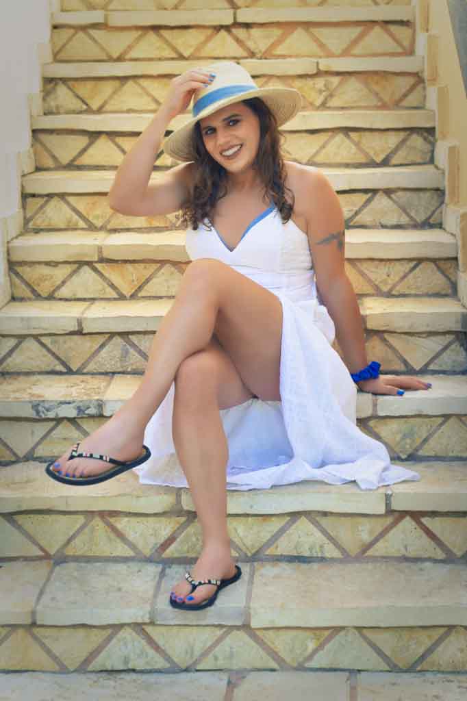 Full body shot of Jessica in a white sun dress, seated on the steps with her hand on her sunhat, Zakynthos