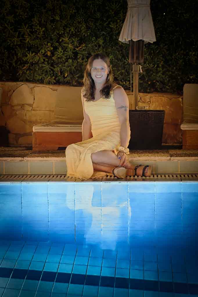 Evening photo of Jessica in a yellow sundress, seated at the side of the pool with her reflection in the blue water, Zakynthos