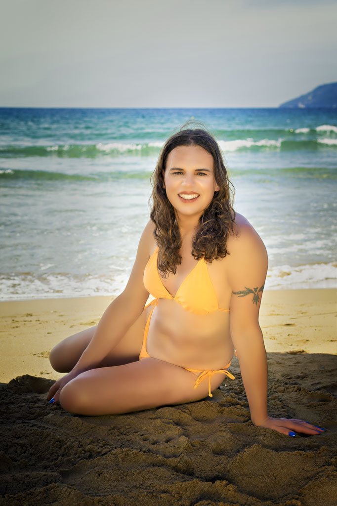 Jessica in an orange bikini, sitting side-on in the shade of a beach umbrella, sea behind her, Gerakas Beach, Zakynthos