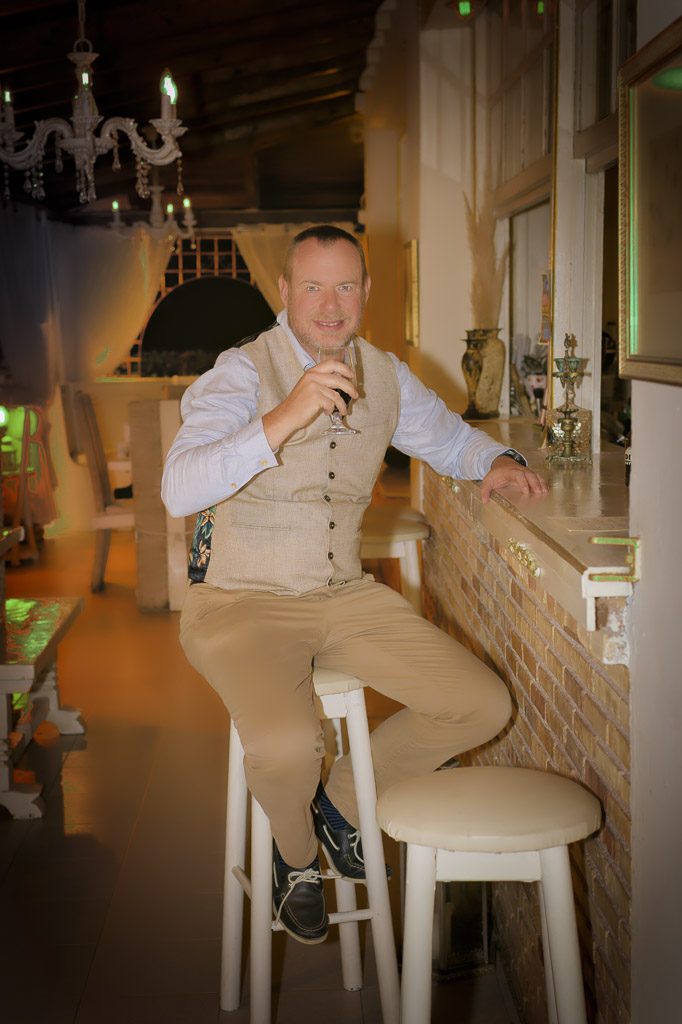 Richard at the hotel bar at night, wearing cream chinos and a cream waistcoat, with a drink, Zakynthos