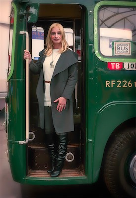 Model in a grey coat and black boots standing in the doorway of a vintage green London bus at a museum