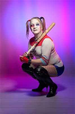 Model in a Harley Quinn-style costume crouching with a baseball bat against a purple and pink backdrop